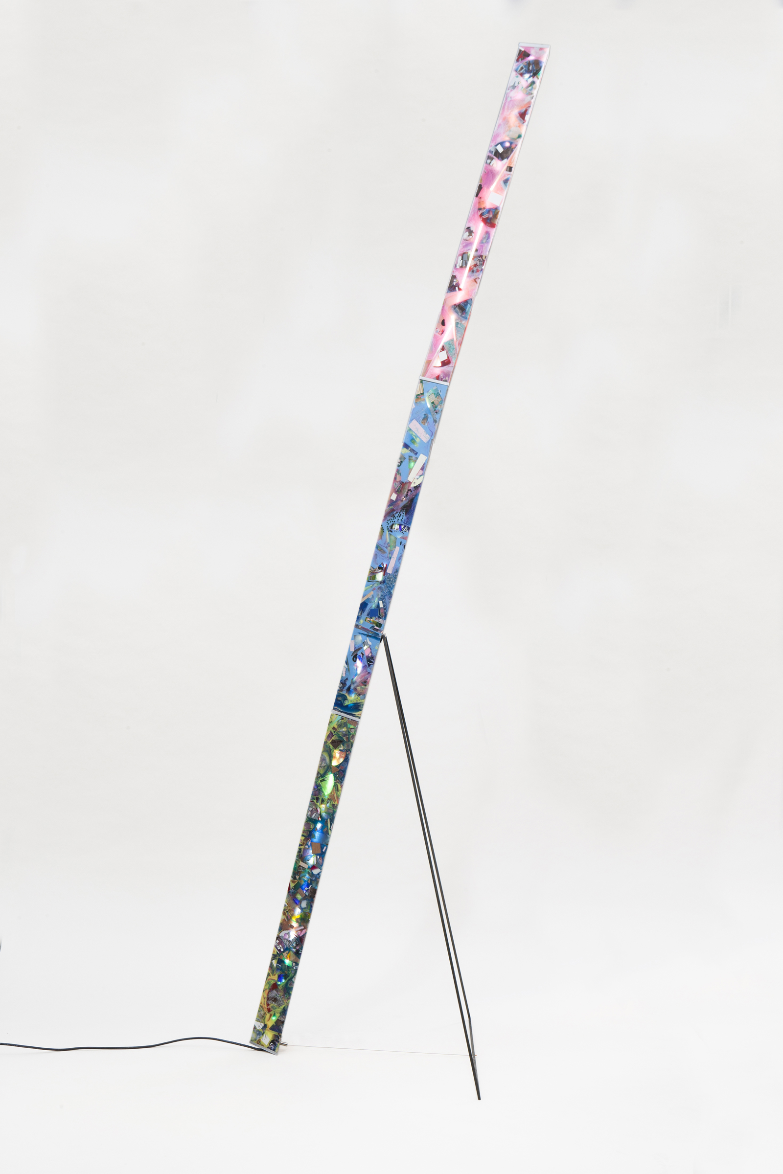 side view of floor lamp consisting of a long pillar of resin of various colors and material inlays, triangular black metal frame and cord.