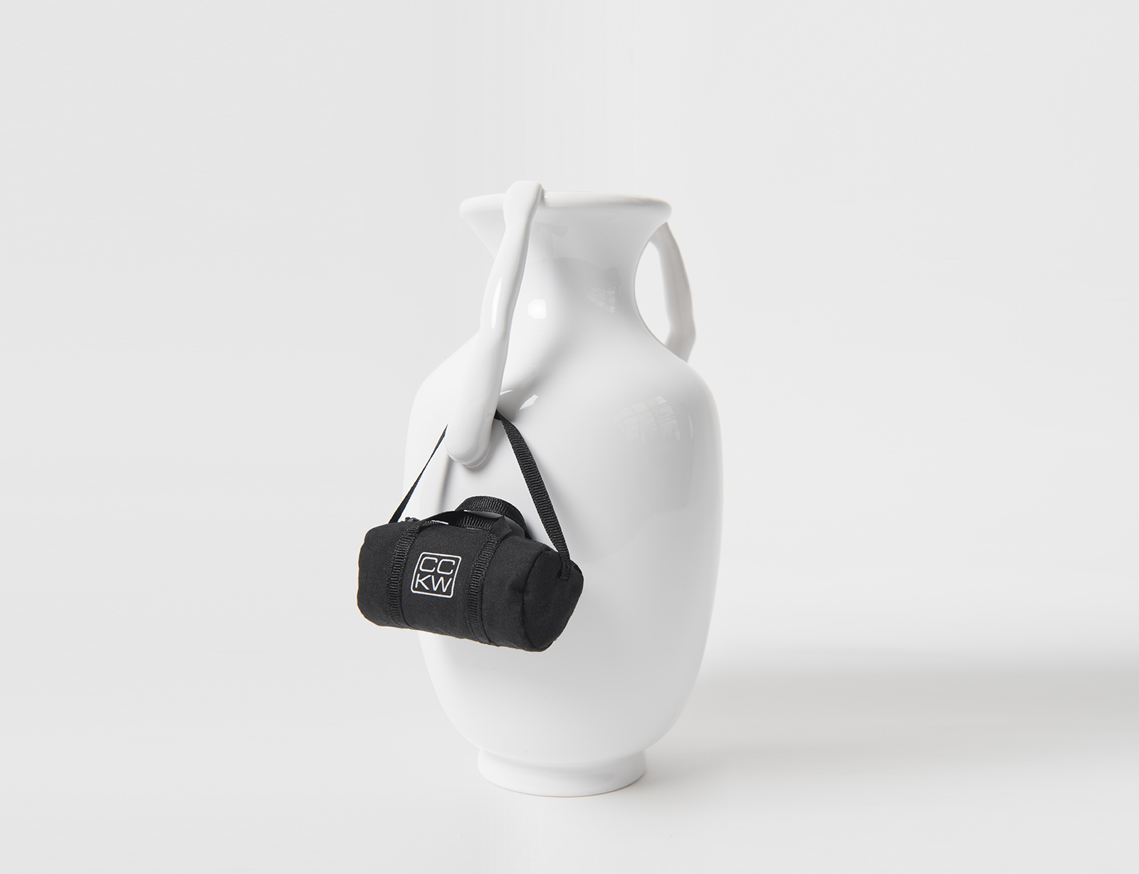 white porcelain vase with two arms extending upward and holding rim of vessel. around one arm is a miniature black duffel bag.