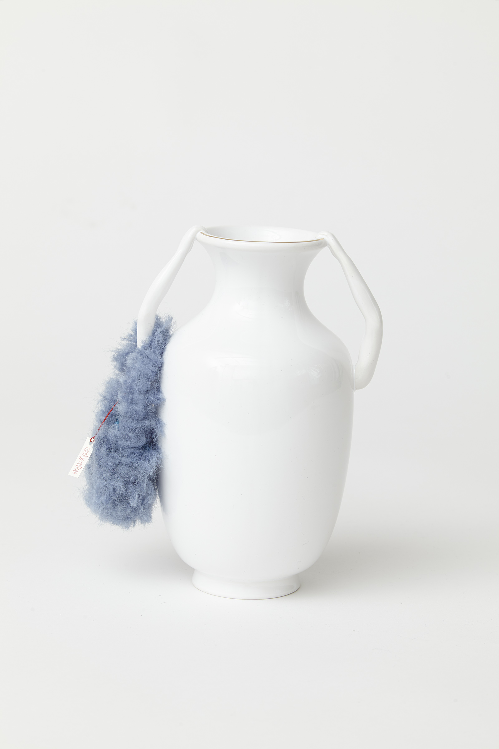 white porcelain vase with two arms extending upward and holding rim of vessel. around one arm is a miniature fuzzy light blue bag