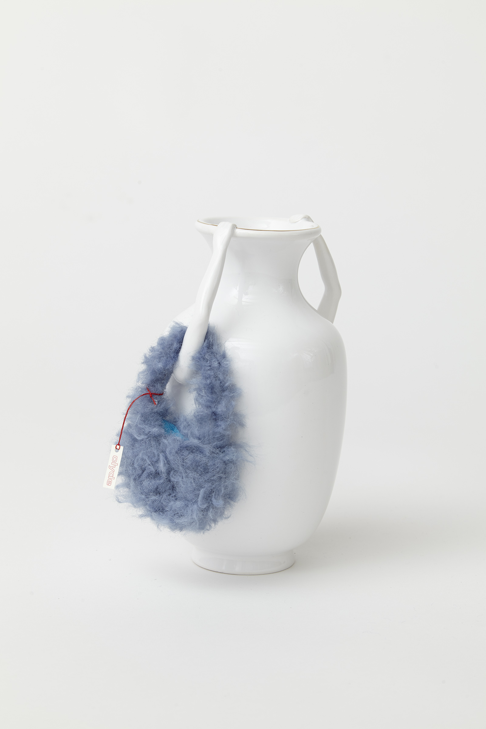 white porcelain vase with two arms extending upward and holding rim of vessel. around one arm is a miniature fuzzy light blue bag